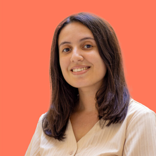 Photo de Lina Djebali, une jeune femme souriante aux cheveux longs et bruns, portant un chemisier clair. Elle se tient devant un fond uni de couleur corail vif.