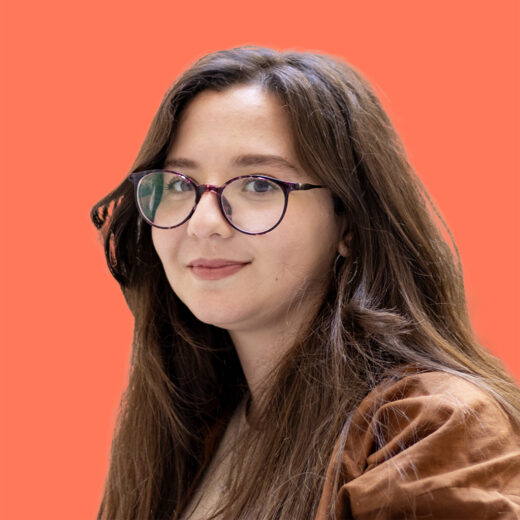 Portrait de Lydia Boussaid, une femme aux cheveux bruns longs et lisses, portant des lunettes rondes à monture marron. Elle a un teint clair, un léger sourire et porte une veste marron. Le fond de l'image est uni et de couleur orange.