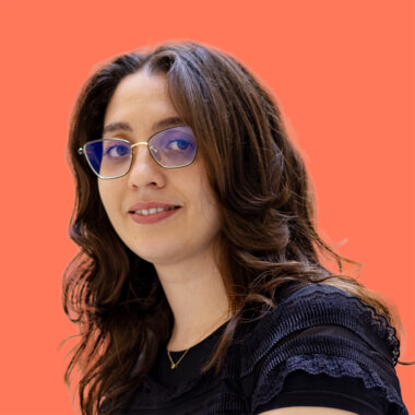 Photo de Sarah Talbi, une jeune femme aux cheveux longs et châtains, portant des lunettes et un haut noir à manches courtes avec des détails en dentelle. Elle sourit légèrement et se tient devant un fond uni de couleur corail vif.